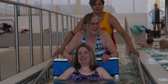 woman in a pool wheelchair with a woman pushing it into the water with lifeguard behind them