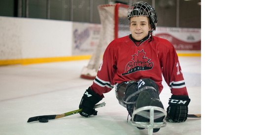 young kid sitting on a sledge in hockey gear with hockey net behind