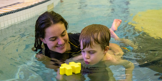 Instructor holding child in water while child blows a block across the water in a public pool