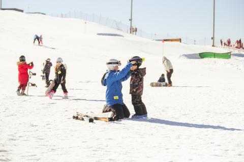 parent putting a helmet on a child on ski hill with skis and kids with snowboards behind